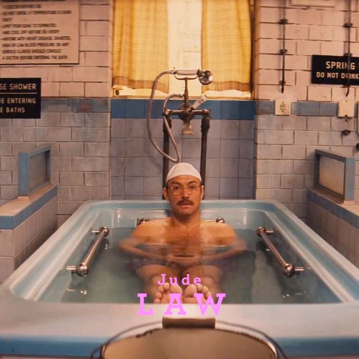 a man in a white bathing cap is relaxing in a bathtub full of water. he is in a tiled bathroom, and the tub is white with blue trim. the man is smiling, and the tub is filled with water. the man is wearing a white bathing cap and a white towel around his waist. the bathroom has a shower, and the walls are tiled with white and blue tiles.