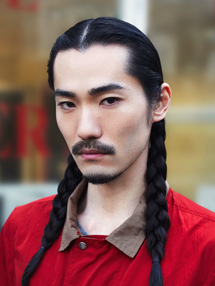 a portrait of an asian man with black hair parted in the middle, styled into two braids. he has a moustache and a short beard. he is wearing a red shirt with a collared neck. the background is blurred.