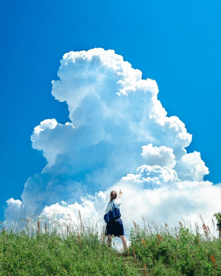 a woman with a backpack stands on a grassy hill, pointing up at a large, fluffy cumulus cloud in a bright blue sky.