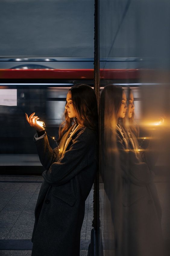 a young woman stands on a subway platform at night, her profile illuminated by the warm glow of string lights. she is leaning against a wall, her reflection mirrored in the glass beside her. a red train streaks past in the background, blurring with speed.