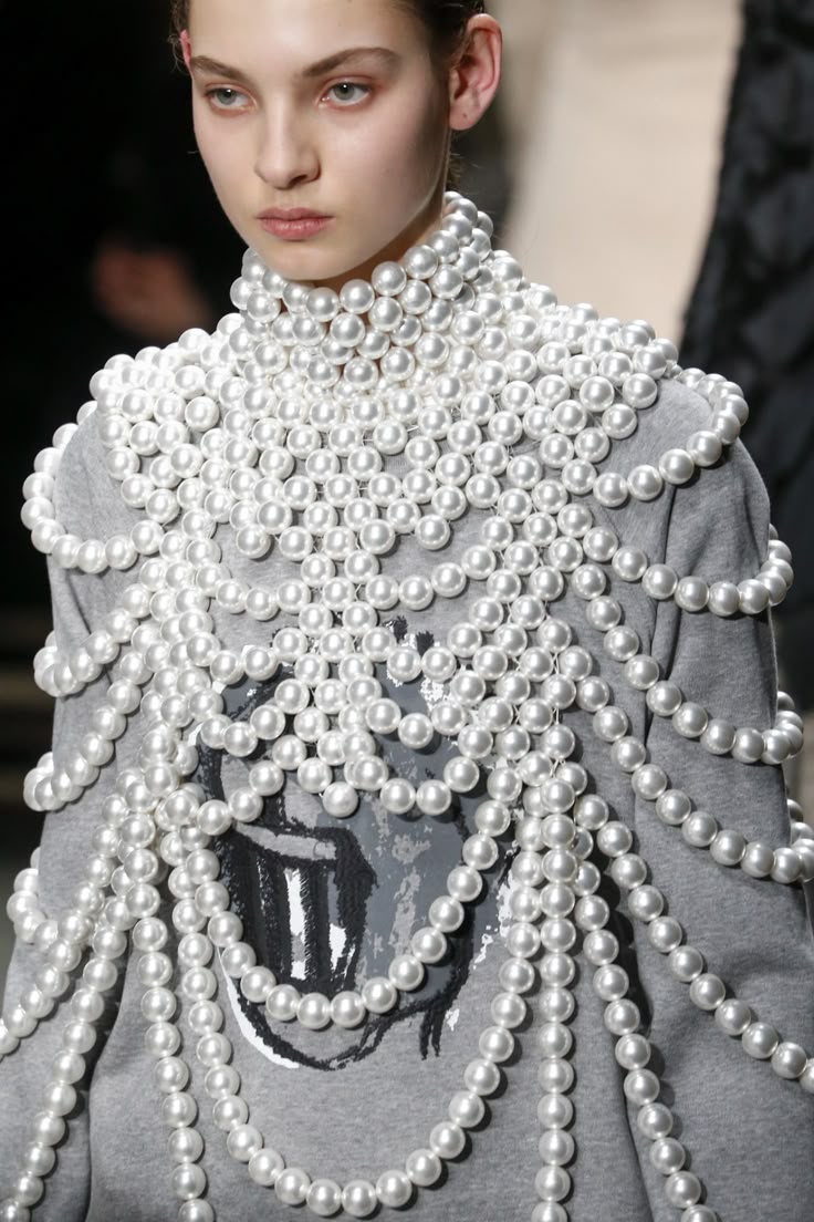 a model on a runway wearing a grey sweatshirt heavily adorned with multiple strands of white pearls. the pearls form a high collar and drape down the front and shoulders of the sweatshirt. the sweatshirt features a graphic design, possibly a stylized image. the model has a serious expression and fair skin.