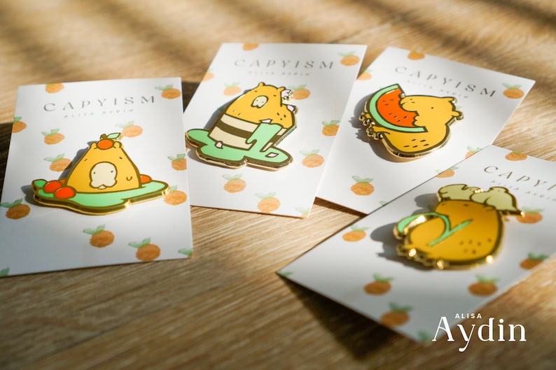 four enamel pins featuring capybaras with different food items are displayed on cards with an orange pattern. the pins show the capybaras with a watermelon, orange, kiwi, and mango. the cards are labeled with "capyism" and "alisa aydin".