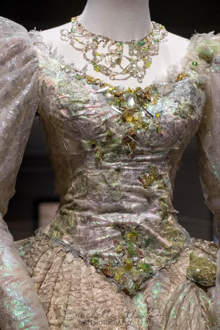 a closeup shot of a mannequin wearing an elaborate dress with a matching necklace. the dress is highly ornamented with beads, sequins, lace, and other embellishments. it features puffed sleeves and a fitted bodice that flows into a full skirt. the necklace complements the dress, with a similar bead and gem design. the overall style suggests formal wear with a focus on intricate detailing and a touch of iridescence.