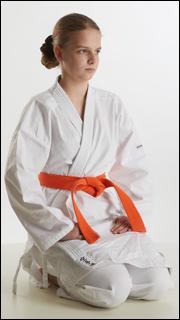 a young woman in a white karate uniform, tied with an orange belt, kneels on the floor in a studio setting. she looks serious and determined. the background is plain white, and the lighting is bright and even.