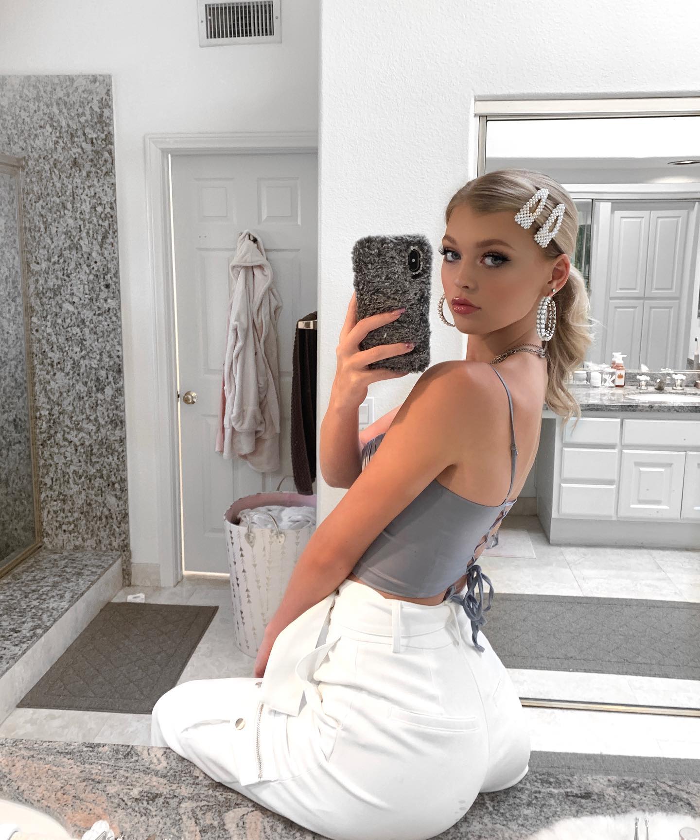 a woman takes a selfie in a bathroom. she is wearing a grey crop top, white pants, hoop earrings, and hair clips. her hair is styled in a messy ponytail. she is holding a phone with a furry phone case. the bathroom has a mirror, vanity, and a door with clothing hanging behind it.