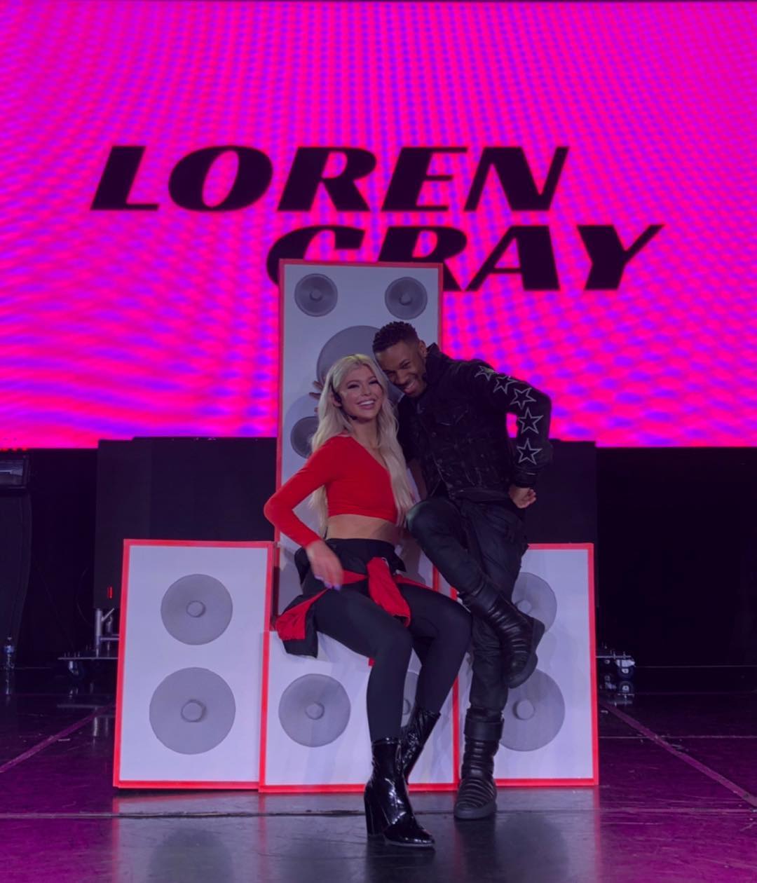 a photo of loren gray and a man on a stage with the words "loren gray" in big bold letters behind them. loren gray is wearing a red top, black leggings, and black boots. the man is wearing a black jacket with stars on the sleeves, black pants, and black boots. they are standing in front of two large speakers.