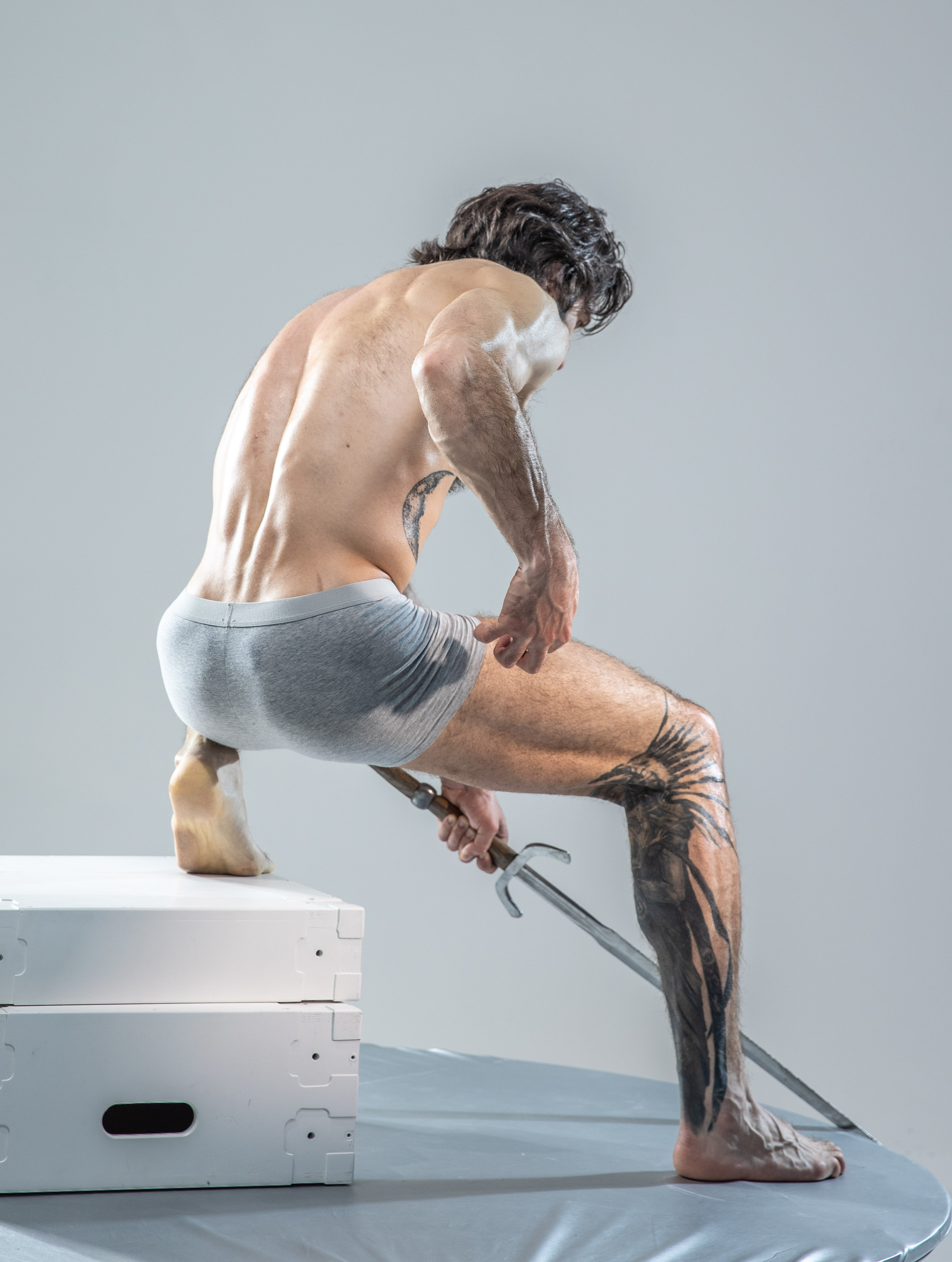 the image showcases a muscular man with tattoos, partially covered in white markings, adopting a dynamic crouching pose on white boxes. he wields a sword with a focused expression, captured from a rear perspective against a minimalist gray backdrop in a studio setting.
