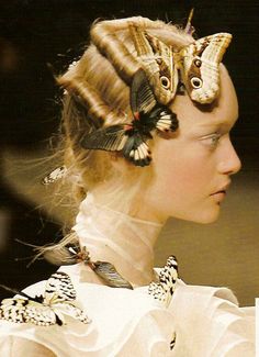 a beautiful blonde woman is wearing a white dress and a very intricate hairstyle adorned with butterflies. her look is whimsical, elegant, and glamorous. the butterflies are placed strategically around her hair and face, giving the overall look a delicate and ethereal feel.
