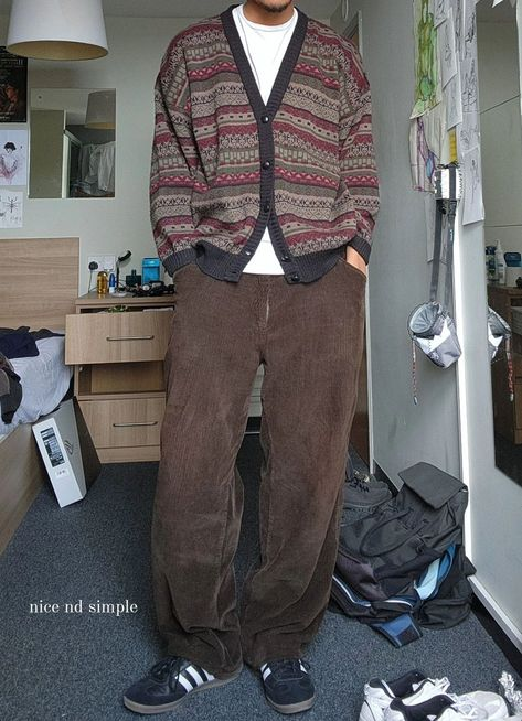 a man standing in a room wearing a brown corduroy pants, a white shirt, and a patterned cardigan. he is wearing sneakers and has a backpack on the floor next to him.