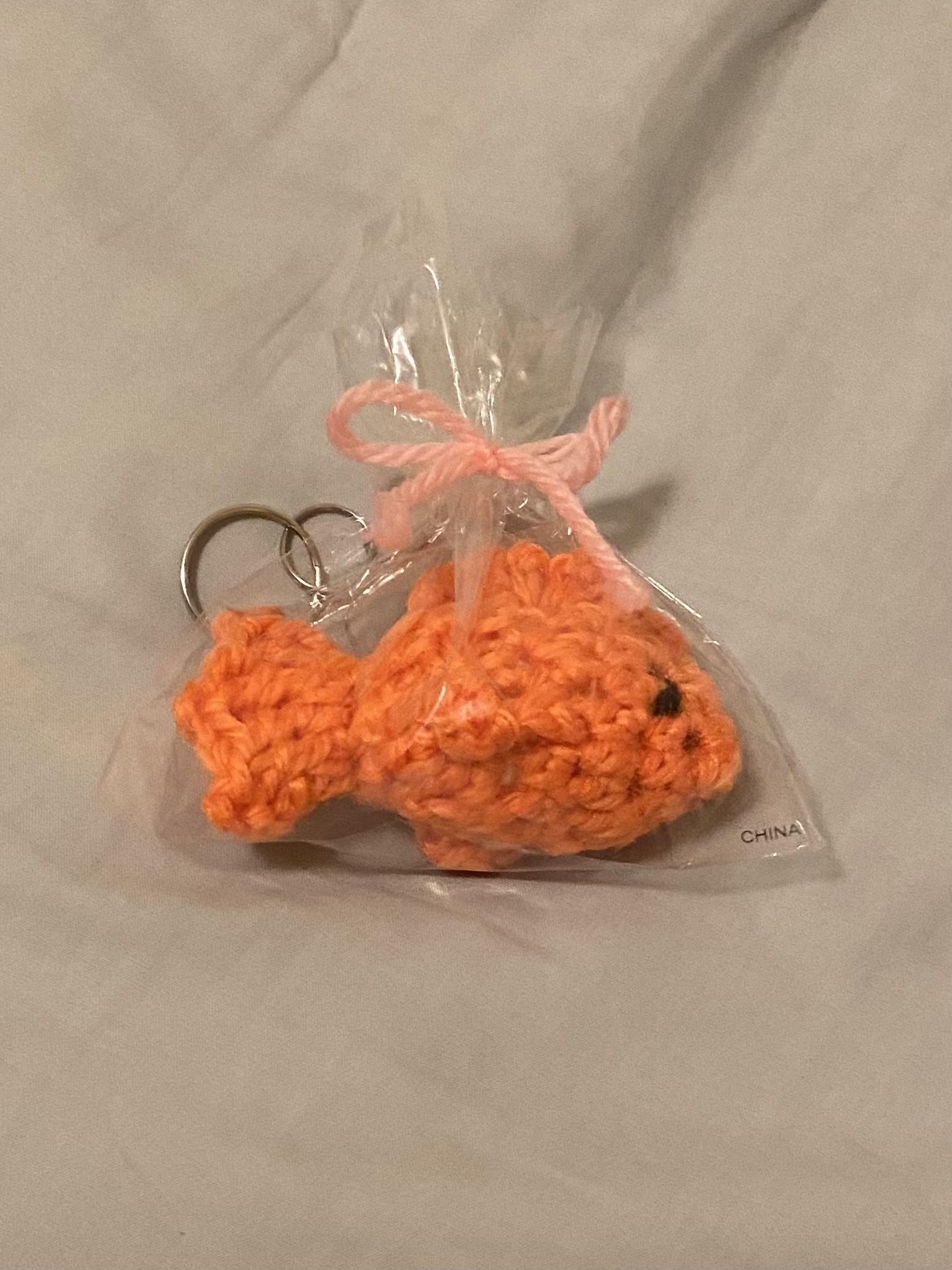 a closeup shot of a small, crocheted orange goldfish keychain in a clear plastic bag tied with a pink ribbon. the keychain is handmade and appears to be a small, cute toy or accessory. the bag has a "china" label on it.  the background is a light beige fabric.