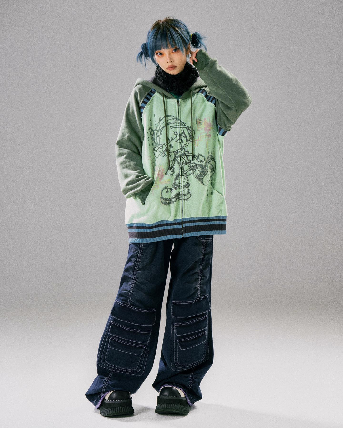 a full shot of a young woman modeling a pastel green oversized hoodie with an animeinspired graphic print. the hoodie has a contrasting dark blue and light blue stripe detail at the bottom and cuffs.  she is wearing dark blue wideleg cargo pants with multiple pockets and chunky platform shoes. her hair is styled with light blue strands pulled back in halfponytails.  the background is a muted grey. the overall style is streetwearinspired with an anime/cartoon graphic element.