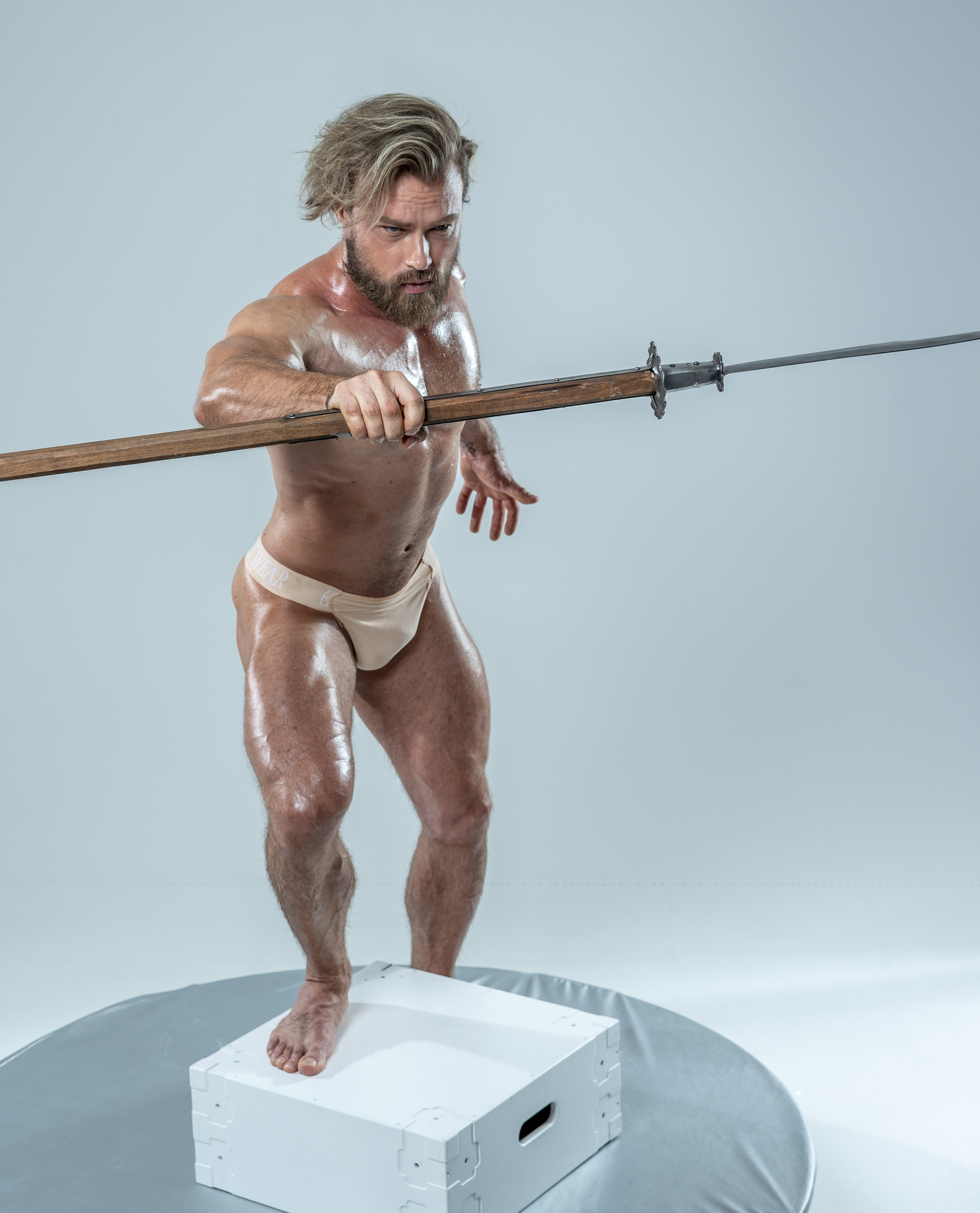 a muscular man with long blonde hair in his underwear is holding a spear. he is standing on a white cube with a grey backdrop behind him. the image is welllit with soft lighting.