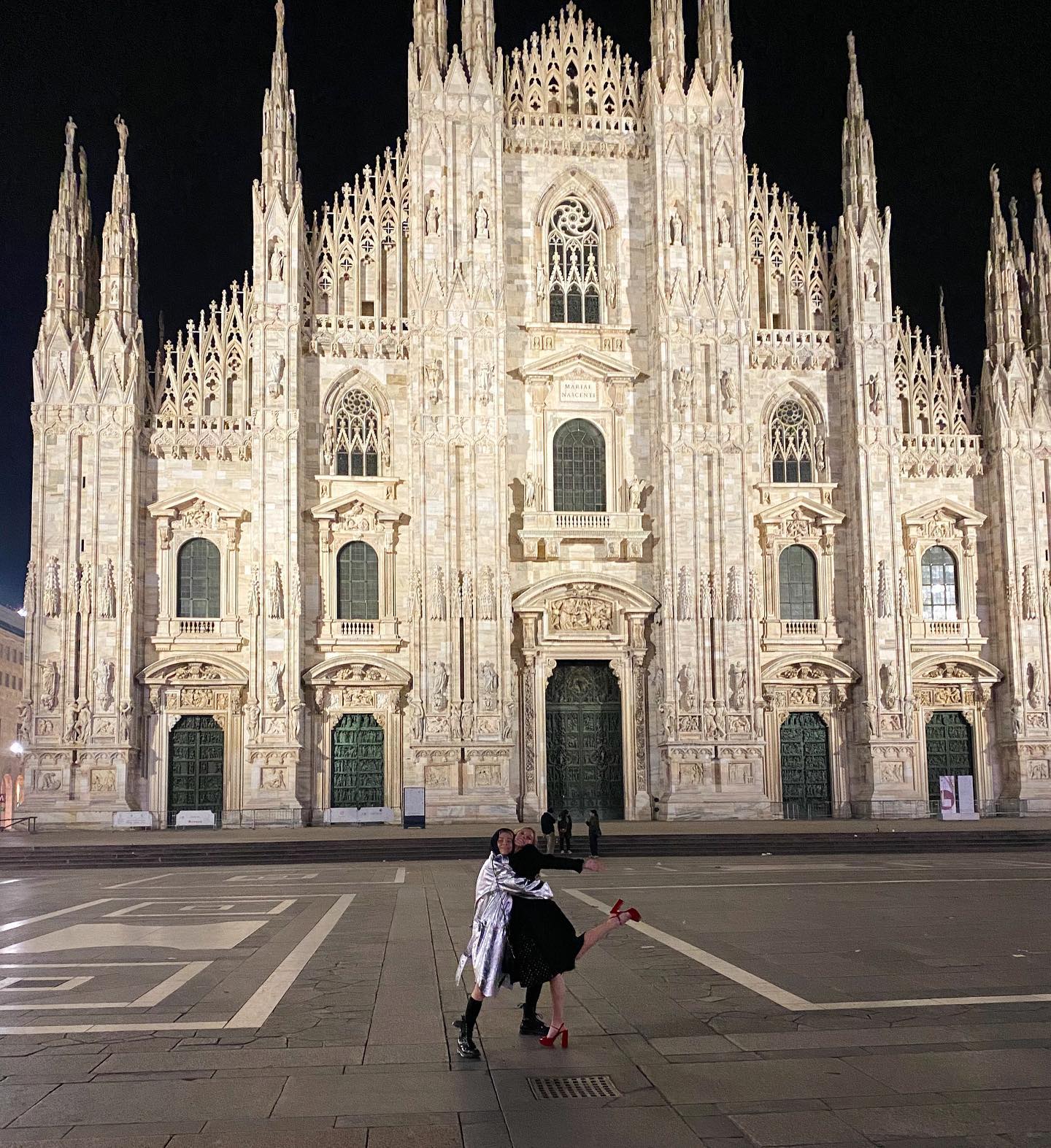 a nighttime photograph of the duomo di milano (milan cathedral) in italy.  two women are hugging in the foreground of the piazza in front of the cathedral.  the cathedral is beautifully lit up, showcasing its intricate gothic architecture. the women appear happy and are dressed in stylish outfits. the overall scene is one of travel, tourism, and a joyous moment captured in a stunning urban setting.