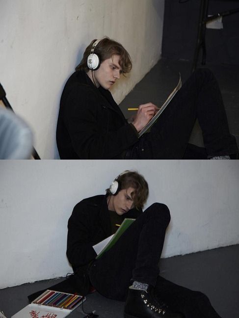 the image features a young man engrossed in his artwork. he is seated on the floor, wearing headphones and deeply focused on the drawing in his sketchbook. the environment appears to be an art studio, with soft, natural light illuminating the scene.  the overall aesthetic is contemplative and artistic, emphasizing the man's creative process and solitude.