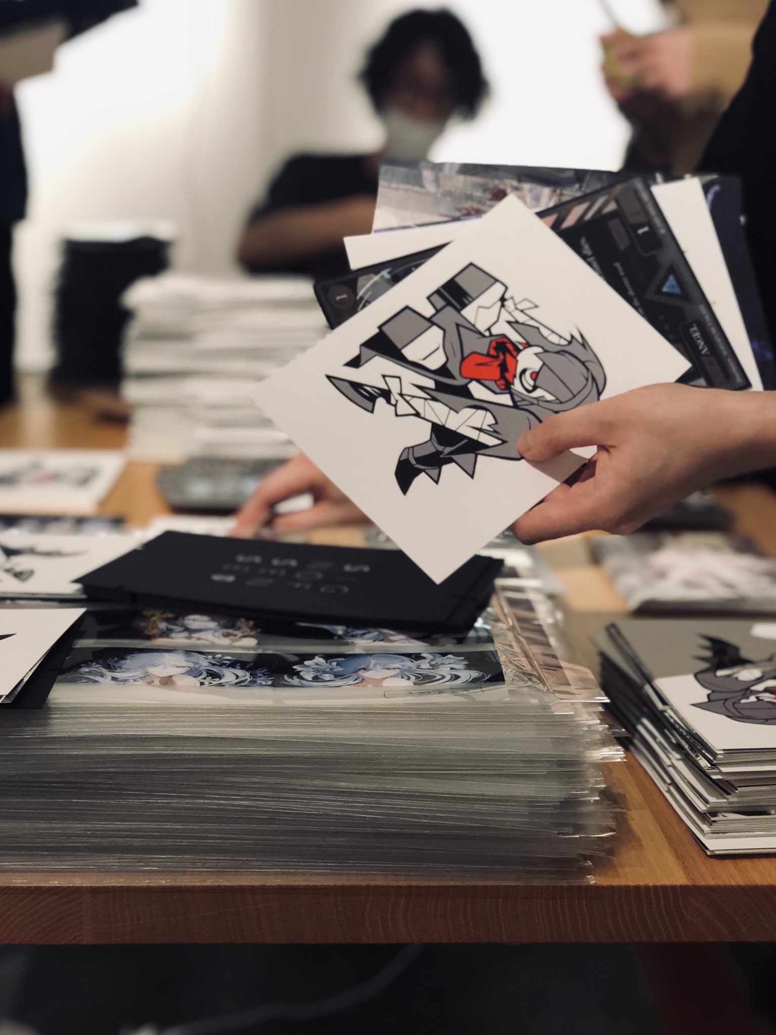 a person is holding up a print of an animestyle illustration featuring a character in a dynamic pose. the print is in black and white with red accents, and they are surrounded by stacks of similar prints and merchandise, suggesting they are at an artist alley or similar event. the overall lighting is bright, likely from overhead fluorescent lights, creating a busy and bustling atmosphere.