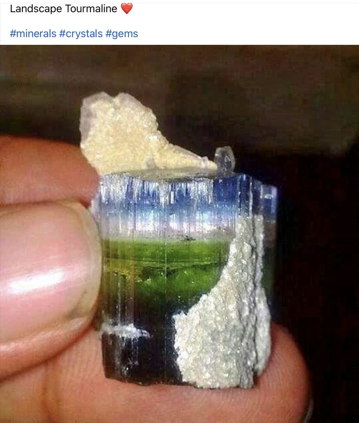 a closeup shot of a landscape tourmaline crystal being held in a person's hand. the crystal has a clear blue top, a green middle section, and a dark black bottom. there are pieces of white and tan rock attached to the crystal. the image is a social media post with hashtags.