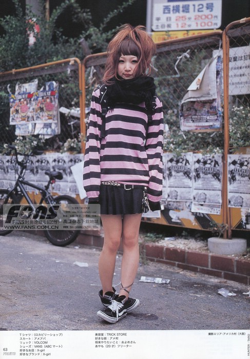 young asian woman is standing on a city sidewalk, posing for a street fashion photo. she is wearing a pink and black striped shirt, a black skirt with a studded belt, black sneakers with white laces, and a black scarf with a skull and crossbones print. she has brown hair with blonde highlights styled in a messy updo and is wearing dark eyeliner. she is standing in front of a chain link fence with posters and flyers on it. there is a bicycle parked on the sidewalk behind her. the photo appears to be from a magazine, as there is japanese text visible.