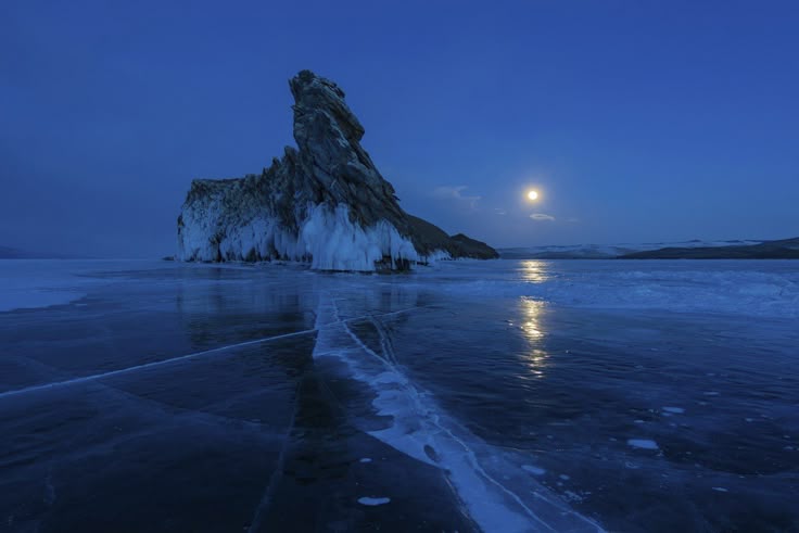 the image showcases a rock formation covered in ice along a shoreline at night, with the moon illuminating the frozen lake. the ice stretches out in the foreground, reflecting the moonlight, while the rock formation stands tall against the dark blue sky. in the background, a distant shoreline is visible. the overall scene captures a serene and icy winter landscape.