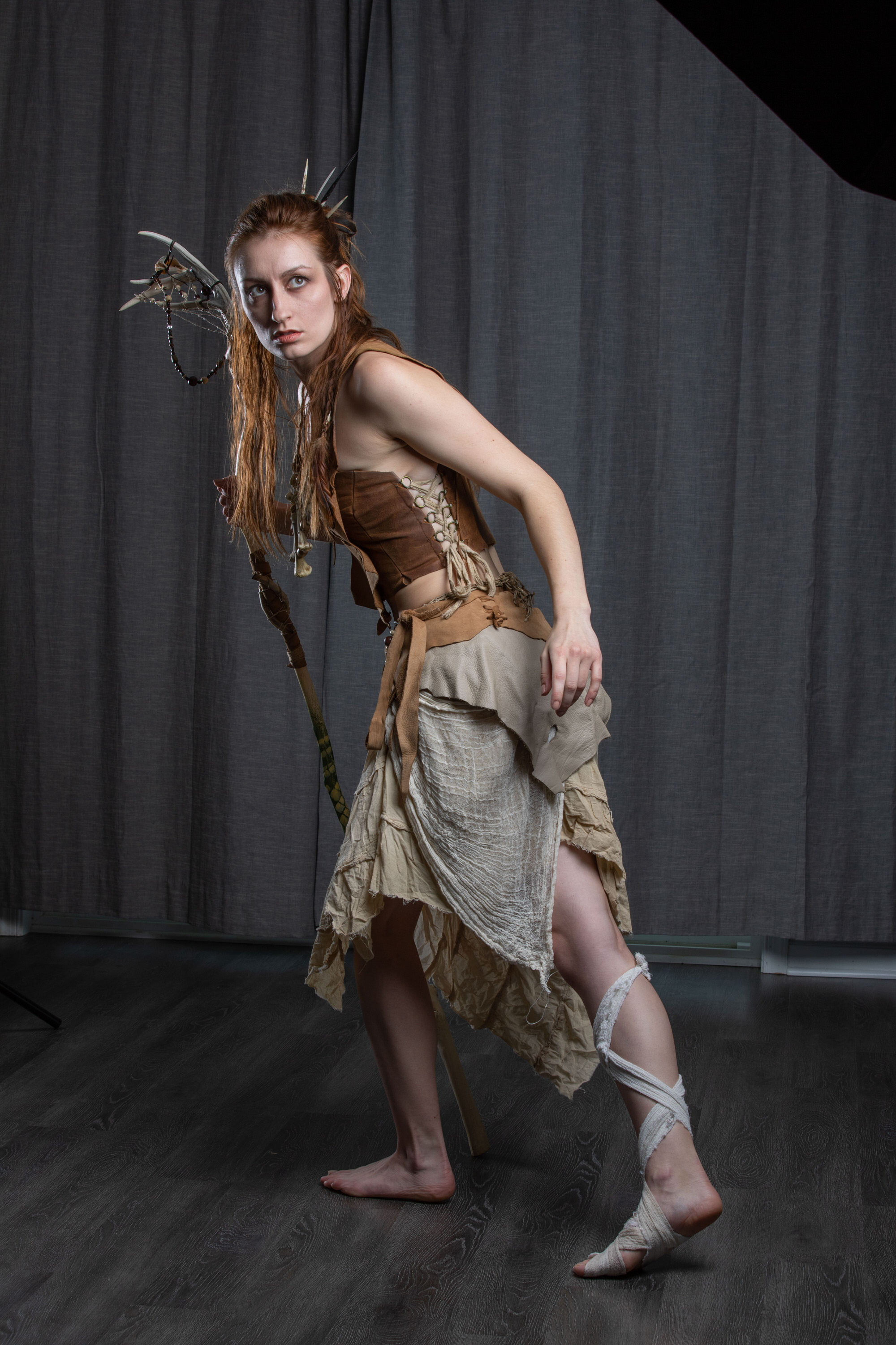 a female model stands in a warrior costume with a staff in a studio setting. she has on tribalinspired clothing and accessories. her costume features earthy tones and natural materials.