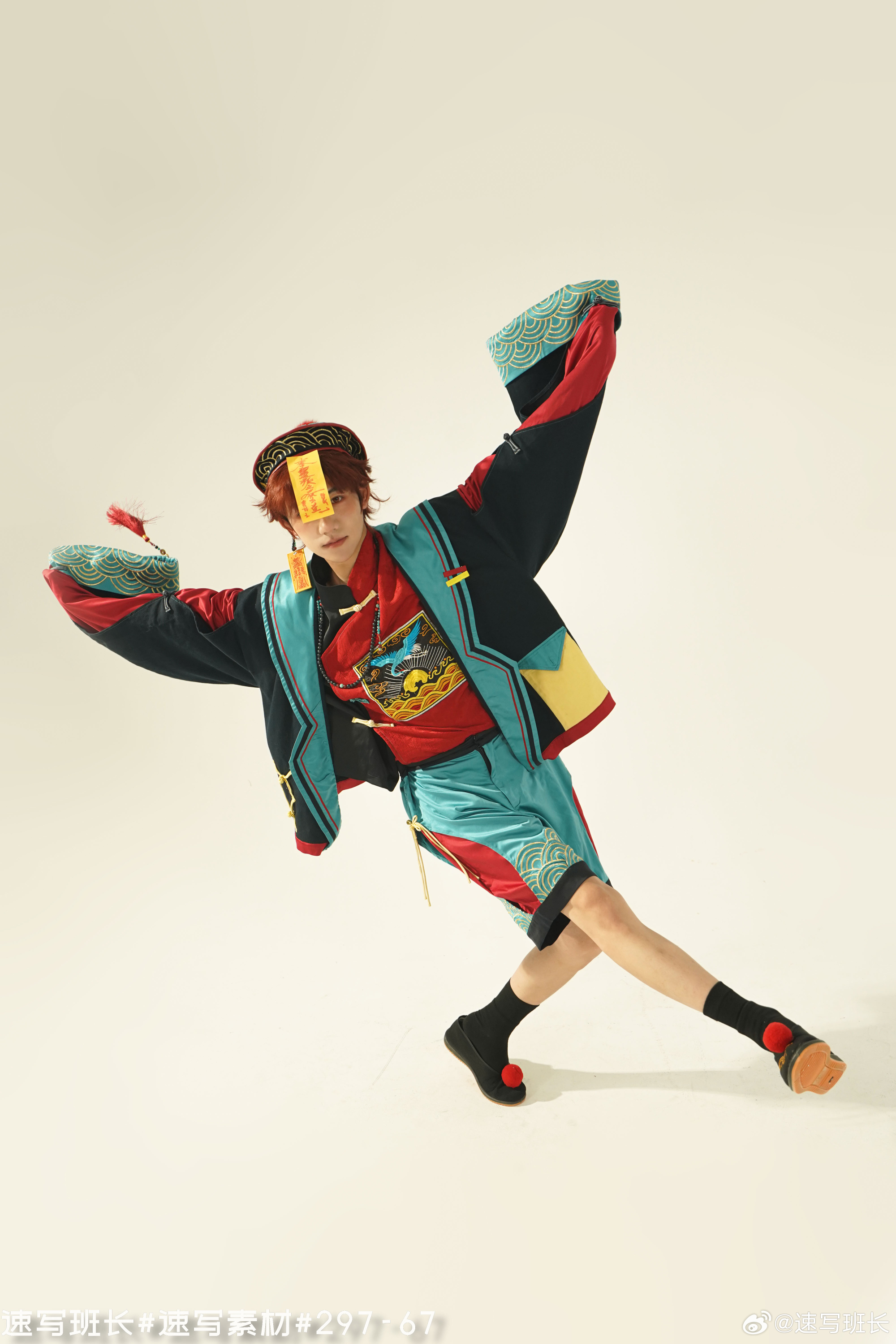 man with reddishbrown hair is captured midpose against a stark white backdrop. he's clad in a vibrant ensemble of traditional chinese attire.  the outfit features a red shirt layered with a blue and yellow jacket, adorned with intricate patterns and chinese characters.  his teal shorts mirror the jacket's design. black slipon shoes complete the look. the man holds a small yellow drum in one hand and a red object, partially obscured by a yellow paper talisman, in the other.  the studio lighting highlights the vibrant colors of his attire and the dynamic energy of his pose.
