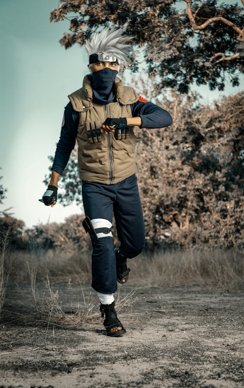a full shot of a person cosplaying as kakashi hatake from the anime naruto. they are running through a dry, grassy field with trees in the background. they are wearing a complete kakashi cosplay, including a grey wig, headband, mask, vest, dark pants with leg guards, and boots. the person is in motion, suggesting a dynamic scene. the overall color palette is muted and earthy tones complement the setting.