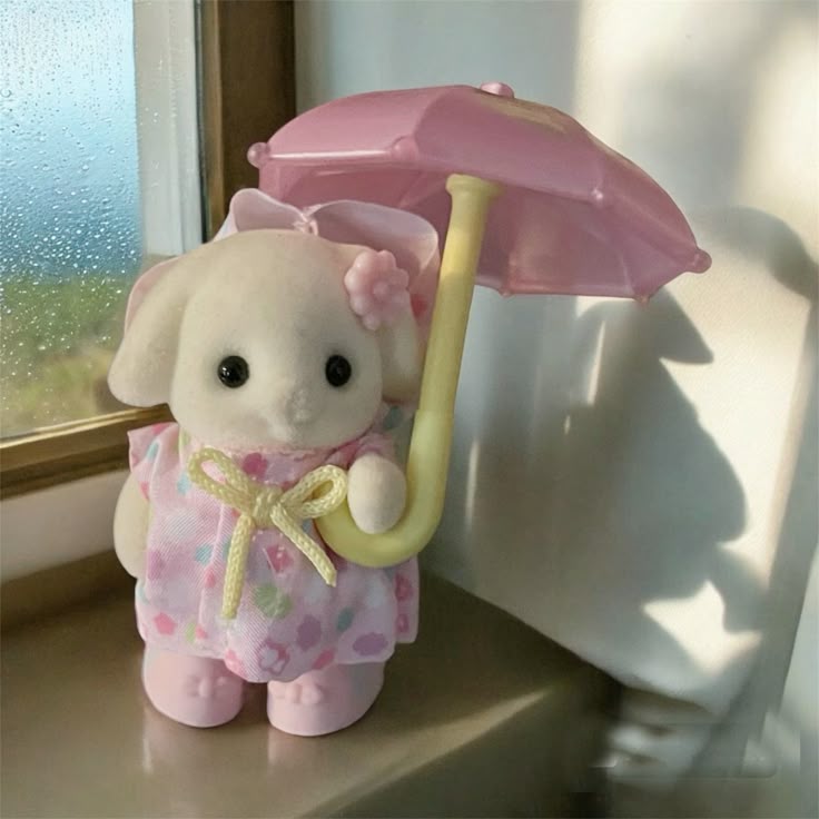 a toy dog figure, presumably a calico critter, stands near a window, holding a pink umbrella. the dog is white and wears a pink floral dress with a yellow ribbon. it has a pink flower and a bow on its head. the umbrella is pink with a yellow handle. the window is visible in the background.