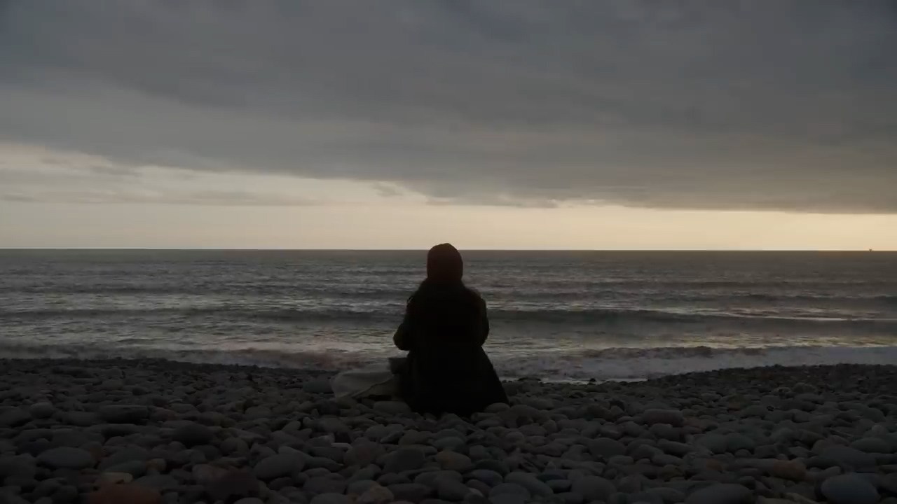 a lone woman sits on a rocky beach facing the ocean as the sun sets behind her, casting her figure in silhouette against the water and sky. the scene evokes a sense of quiet contemplation and solitude.