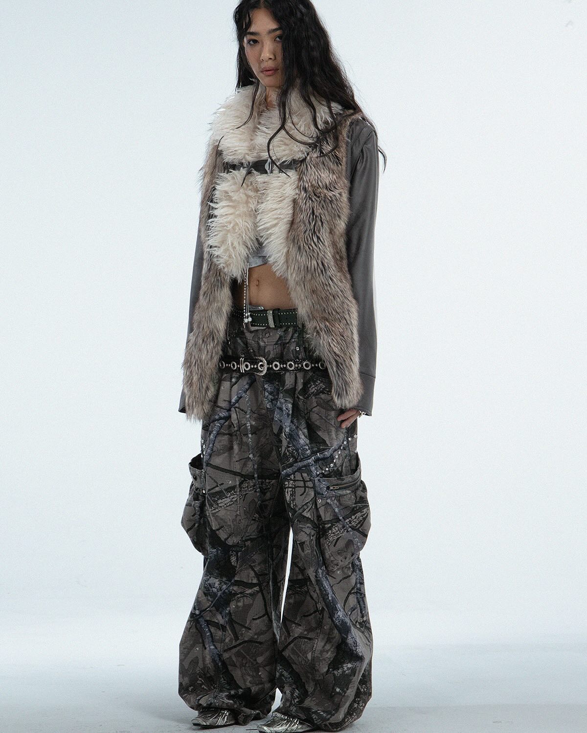 a fulllength studio shot of a female model showcasing a stylish outfit. she's wearing a grey faux fur vest over a grey longsleeved top, paired with camouflage cargo pants and a silver chain belt. the model has long dark hair and is posing against a white background. the overall style is a mix of edgy and wintery elements.