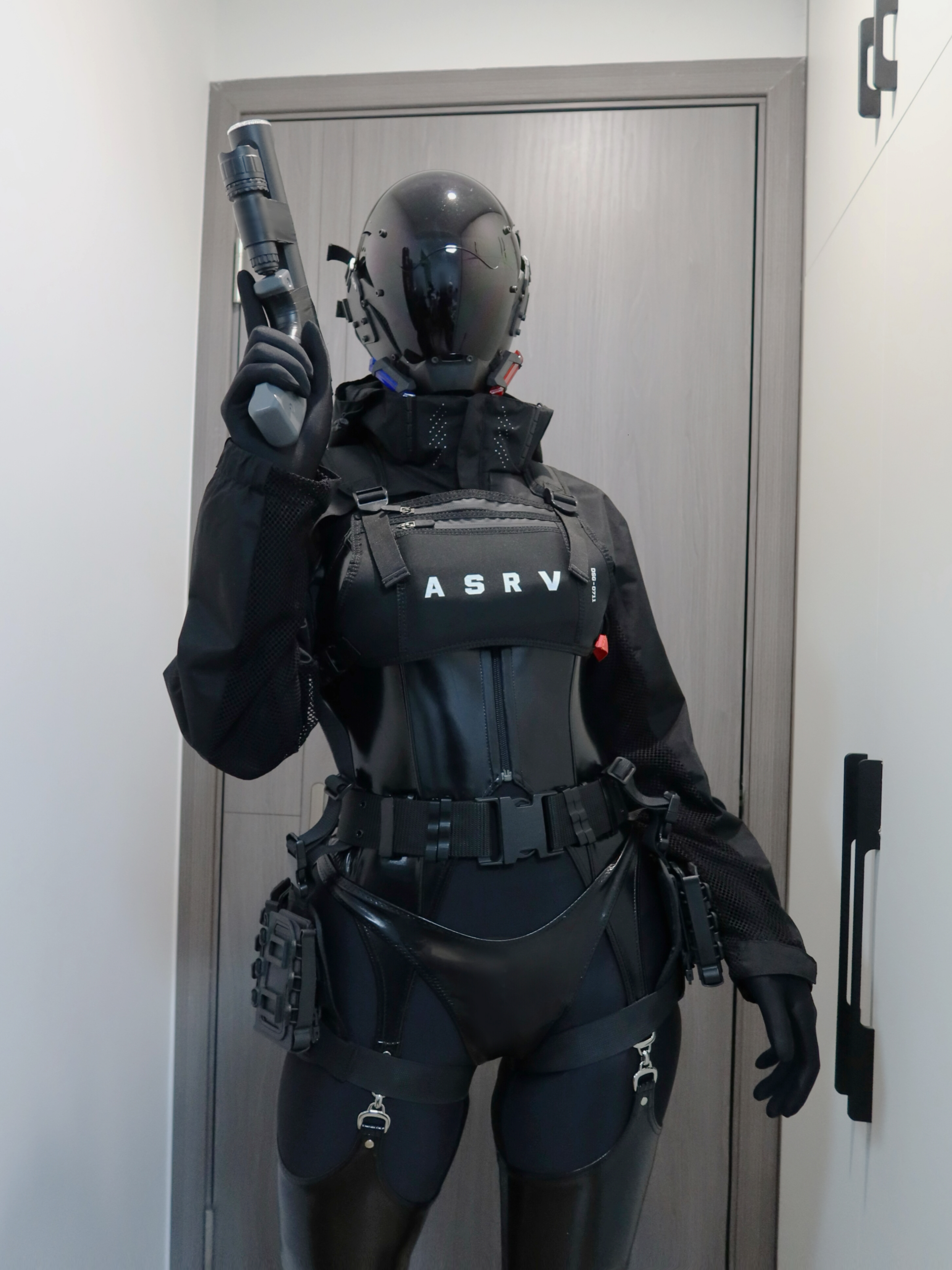 a woman dressed in a black tactical suit with a helmet and a gun in her hand. she is standing in a room with a door and a wall in the background. she is looking to the side of the camera.