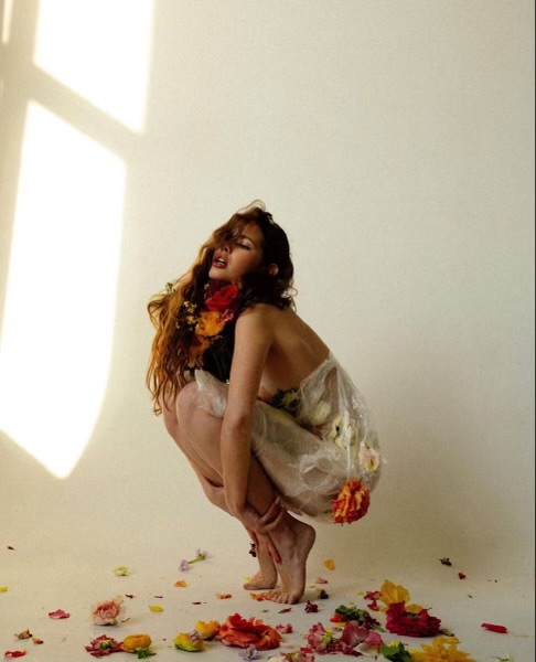 the image shows a young woman with long reddishbrown hair crouching on a light beige floor. she is wearing a sheer, almost transparent, white dress or garment made of what looks like plastic, adorned with various colorful flowers. the flowers are also scattered around her on the floor. the woman's pose is graceful and somewhat sensual, with her eyes closed and head slightly tilted back.  the lighting suggests a studio setting with soft sunlight casting shadows on the wall behind her. the overall aesthetic is artistic and evokes a feeling of delicacy and beauty.
