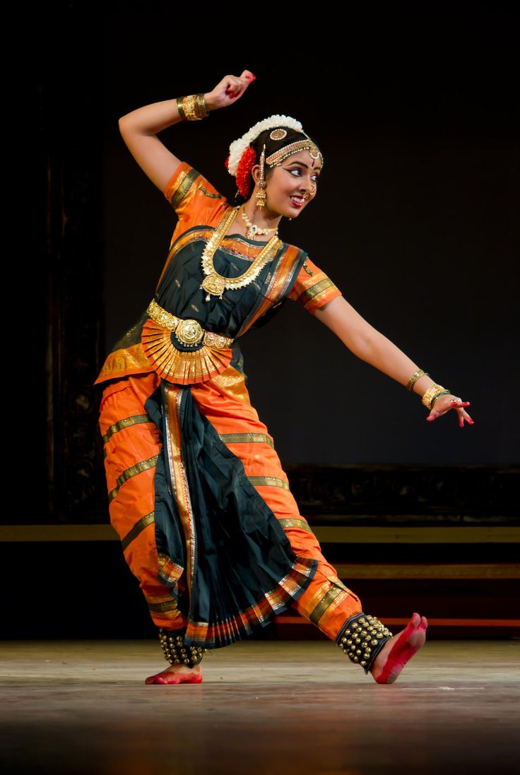 an indian woman in traditional bharatanatyam costume is performing on stage. she is wearing a bright orange and green sari, intricate jewelry, and her face is painted with traditional makeup. she is moving gracefully and with great expression.