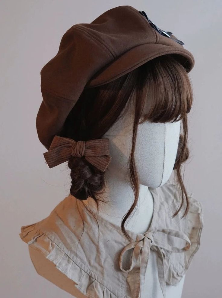 a brown newsboy cap sits atop a brunette wig styled in a braid with a brown bow on a mannequin. the mannequin is wearing a creamcolored top.
