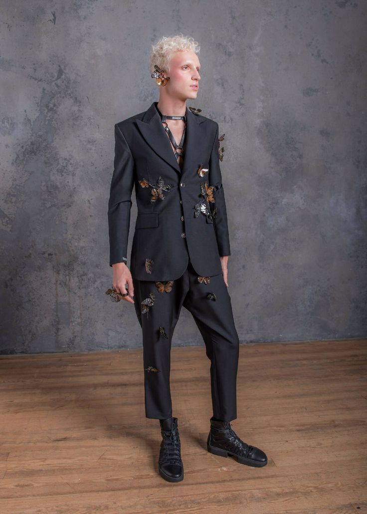 a fullbody shot of a male model showcasing a dark, stylish suit adorned with numerous butterfly accents. the suit consists of a blazer and cropped trousers, both embellished with intricately placed butterflies. the model sports blonde hair, striking earrings, and a subtle necklace. he is posed against a textured grey backdrop, standing on a wooden floor, wearing dark, laced ankle boots. the overall aesthetic is highfashion, blending elements of avantgarde and contemporary styles, with a touch of grunge and edgy flair. the image emphasizes the detail in both the clothing and the model's styling.