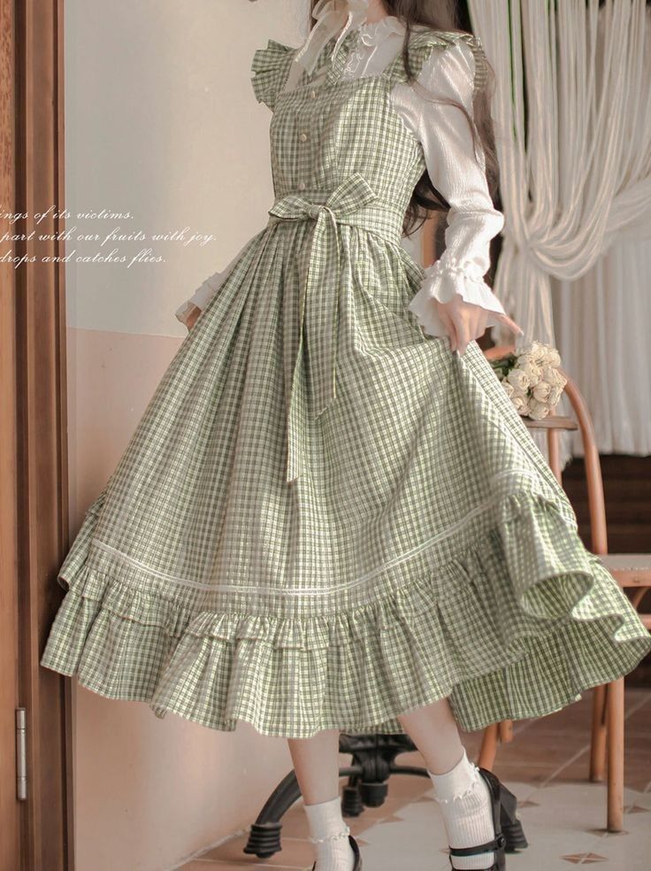 a young woman is wearing a green and white gingham dress with a white blouse underneath. the dress is long and flowy with a ruffled hemline. the woman is standing in a room with a white curtain behind her. she is wearing white socks and black shoes. the lighting is bright and natural.