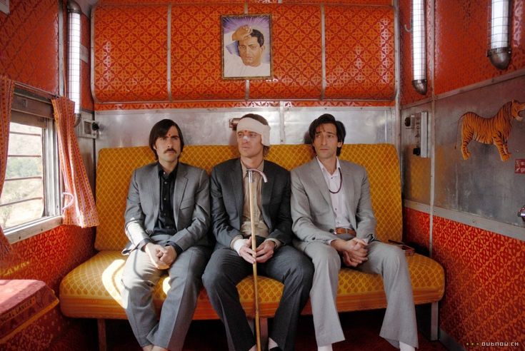 three men are seated on a yellow bench inside an indian train compartment. the men are wearing suits. one of them has a bandage on his head and is holding a wooden cane. the walls of the train compartment are adorned with a red and gold pattern and there is a tiger statue on the wall. the train compartment is lit by overhead lights and a window is visible to the left of the frame.