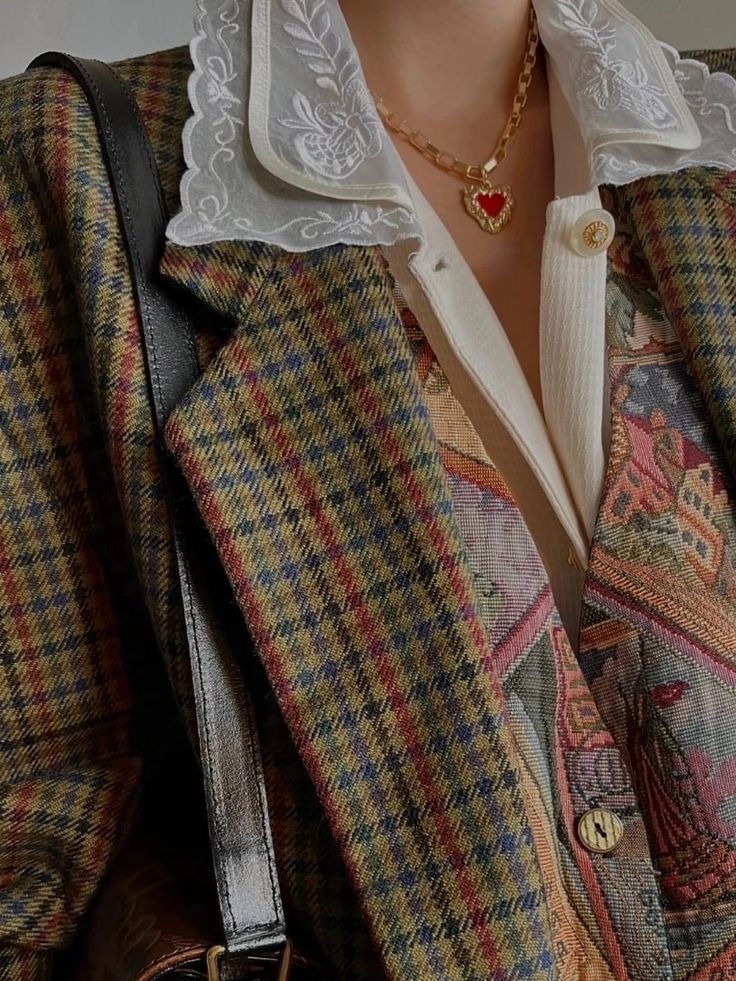 loseup shot of a woman wearing a vintagestyle outfit. the focal point is a brown, green, and red houndstooth tweed blazer layered over a colorful tapestrypatterned waistcoat. the blazer features a delicate white lace collar. the woman also wears a gold necklace with a red heart pendant. the overall aesthetic is retro and autumnal.  a portion of a brown leather bag strap is visible. the image is rich in texture and detail, showcasing the intricate patterns of the fabrics.