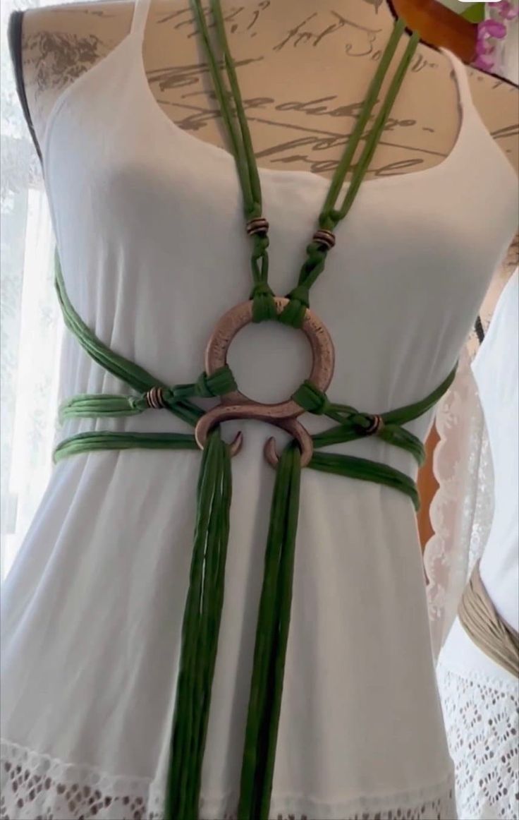closeup view of a handmade green cord necklace styled as a body harness on a white dress. the necklace features coppercolored metal accents and tassels. the dress has a lace trim at the bottom. the image showcases a bohochic fashion aesthetic.  the necklace is worn on a dress form or mannequin.