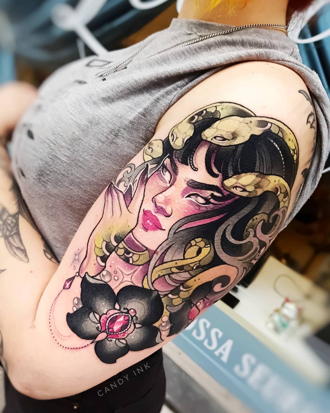 a closeup photograph showcasing a vibrant tattoo of medusa on a person's upper arm. the tattoo, rendered in a neotraditional style, depicts medusa with flowing black hair intertwined with several snakes. her gaze is direct and captivating, while a stylized black flower with a red gem adorns the lower part of the tattoo. the artwork is detailed and rich in color, reflecting the skill of the tattoo artist.