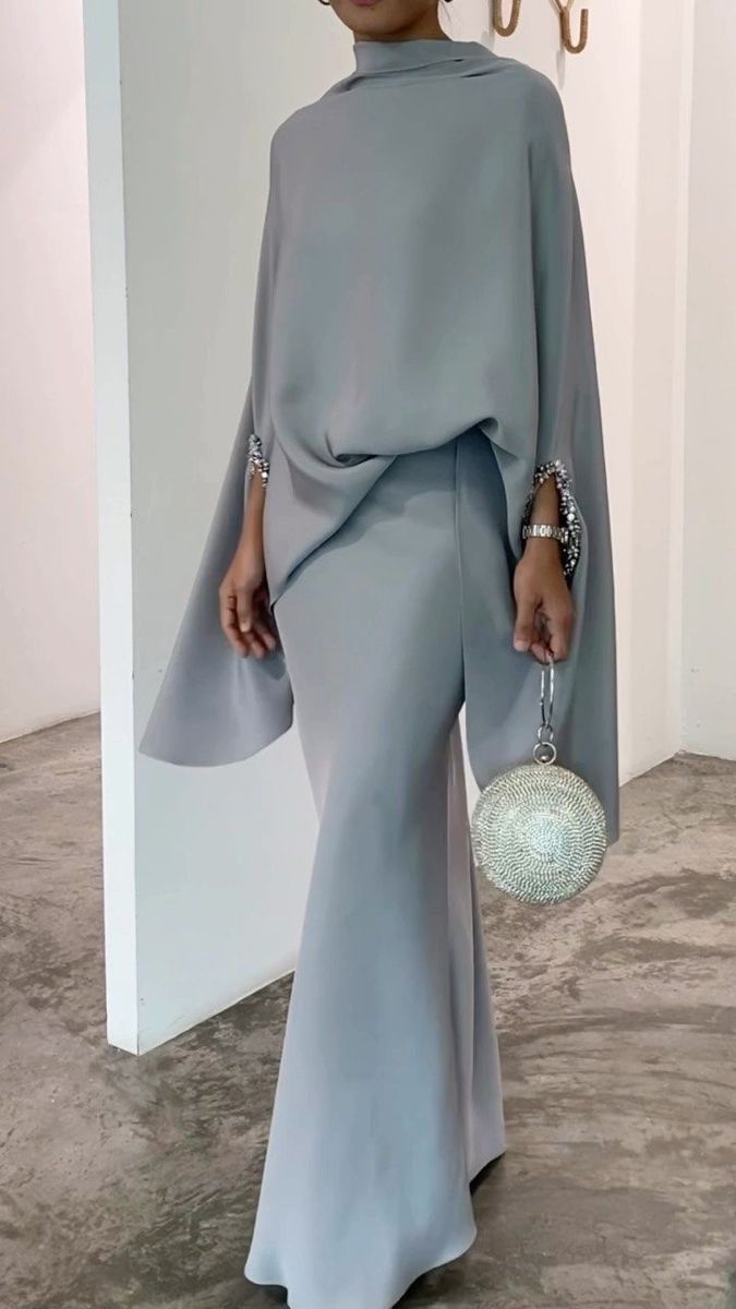 a fulllength shot of a woman wearing a stylish light grey outfit. the outfit consists of a long capelike top and a matching maxi skirt. the cape has a high neck and long sleeves with subtle crystal detailing at the cuffs.  the skirt is formfitting and flows elegantly.  she is holding a small, sparkly clutch bag. the overall style is minimalist, sophisticated, and perfect for a formal event. the image is taken indoors with a clean, simple background.