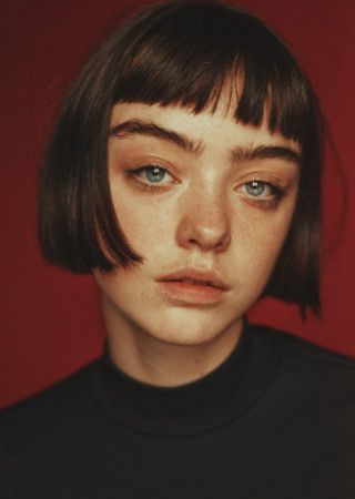closeup portrait of a young woman with a bob haircut and bangs. she has blue eyes and freckles, and her expression is serious and calm.  she is wearing a black turtleneck sweater, and the background is a deep red. the image is styled in a moody, artistic way.