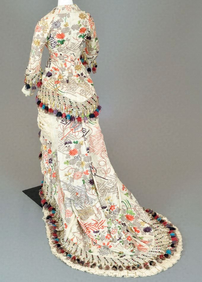 the image shows an antique or vintage gown displayed on a mannequin, viewed from the back. the gown is predominantly offwhite with an elaborate floral and geometric pattern printed or embroidered across the fabric. it has long sleeves and a long train. the gown features heavy embellishments, including tassels and fringe, particularly at the hem and sleeves. the overall design suggests a formal or special occasion dress from a bygone era.