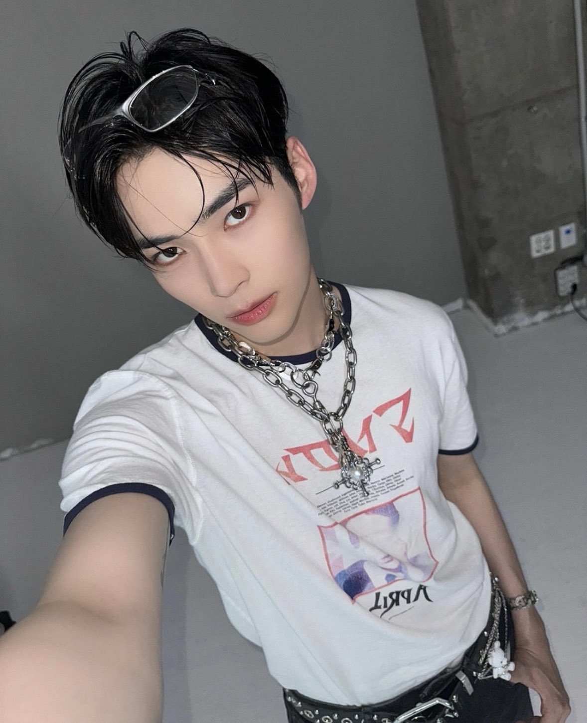 a young man takes a selfie in a studio setting. he's wearing a white tshirt with a graphic print, multiple silver chain necklaces, and black pants with a chain accent. he has short dark hair and is wearing glasses on top of his head. his expression is serious and he's looking directly at the camera. the background is a plain gray wall with some industrial elements visible. the lighting is bright and artificial.