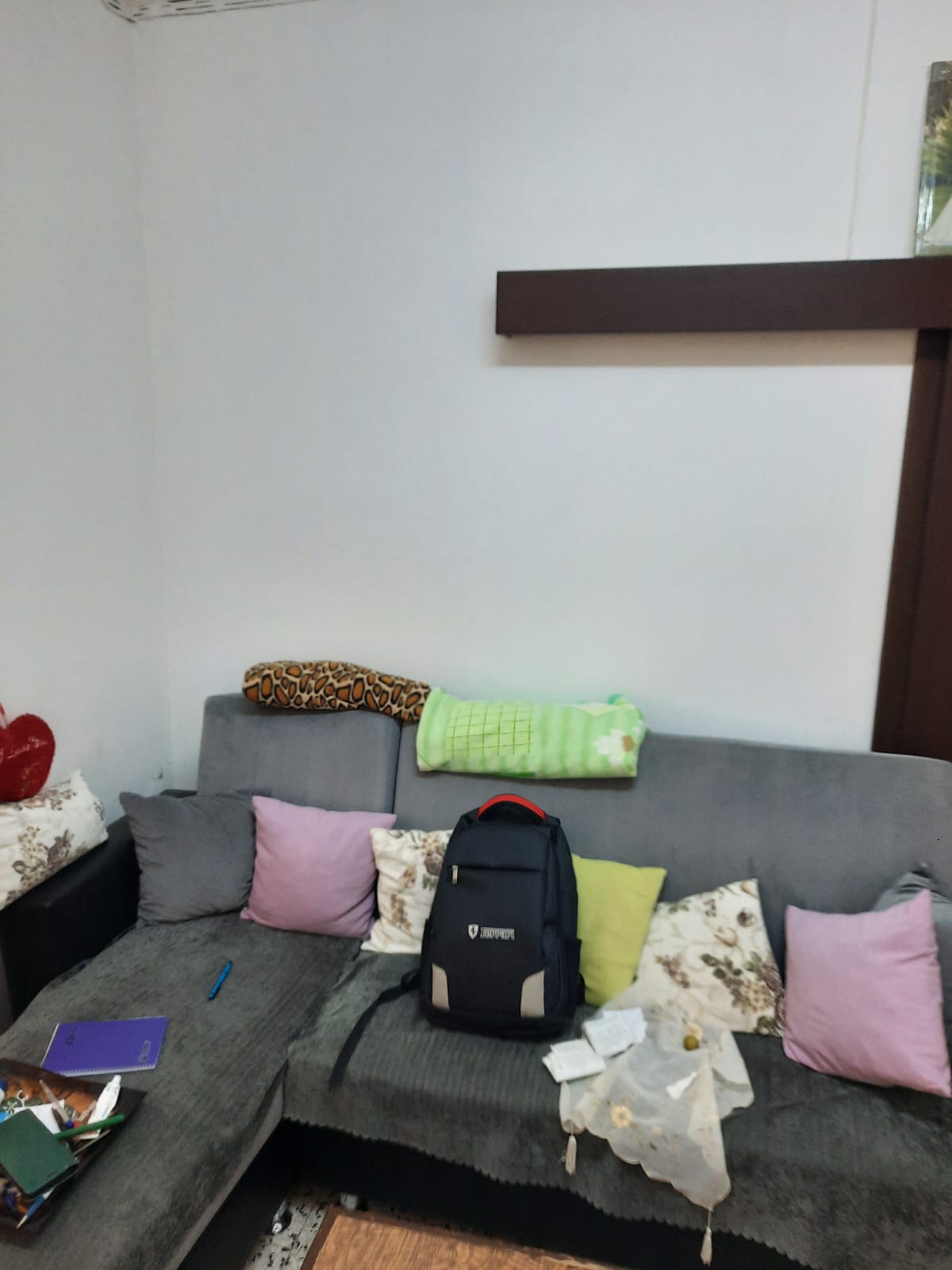 the image shows a living room setting with a gray couch covered in various pillows, a backpack, papers, and other items. there's a small table with a pen, blanket, and additional decorative items in the corner of the couch. the background features a white wall with a dark brown shelf or architectural element.