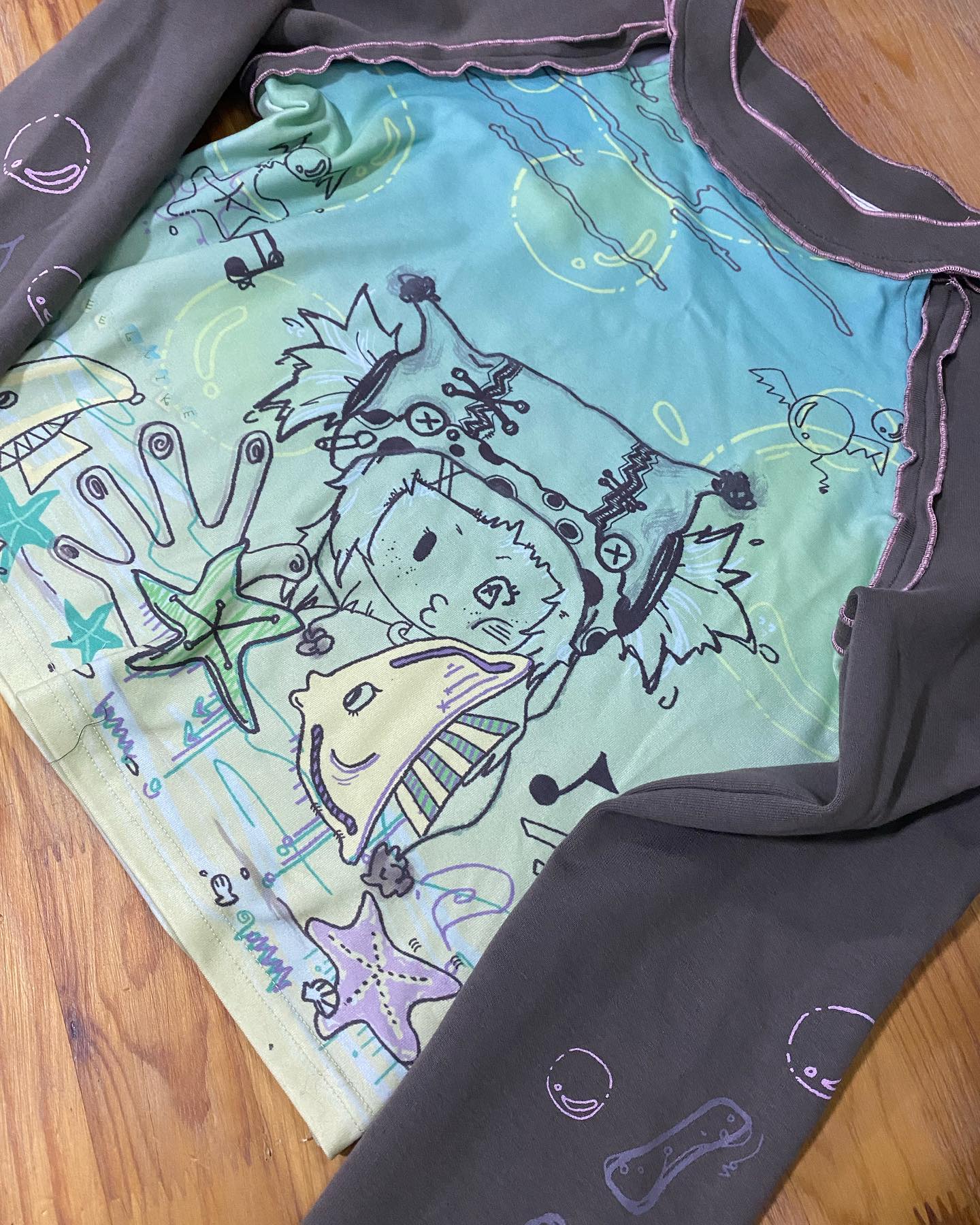loseup view of a cropped, longsleeved shirt featuring a whimsical, handdrawn graphic design. the design incorporates pastel colors, sea creatures (such as starfish and a stylized fish), stars, and other abstract doodles, giving it a cartoonish and playful aesthetic. the sleeves are a darker gray tone compared to the lighter pastel colors of the shirt itself. the shirt appears to be made of a soft, potentially cotton or cotton blend material. the background shows a wooden surface where the shirt is lying.