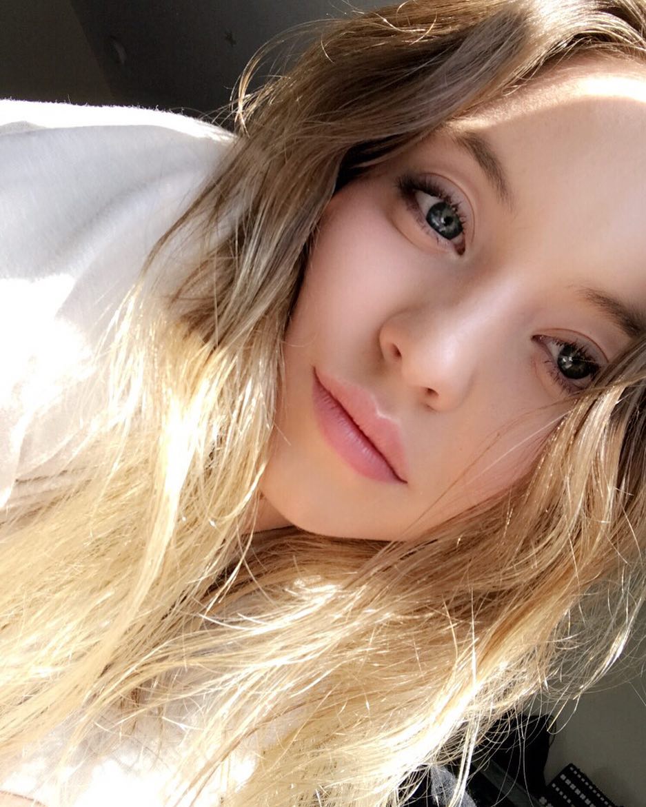 the image is a closeup selfie of a young woman with long, blonde hair.  her hair is somewhat messy and appears to be lit by natural sunlight streaming in from the side. she has fair skin and blue eyes.  she is wearing light makeup and a simple, offwhite top. the background is blurry and indistinct, focusing attention on the woman's face and hair. the overall mood is casual and intimate, suggesting a candid selfportrait.