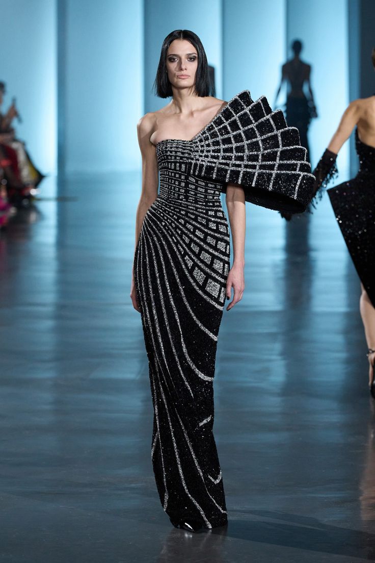 a model is walking the runway wearing a black, oneshoulder evening gown. the gown is heavily beaded and sequined with a grid pattern on the bodice and radiating lines on the skirt. the dress also has a structured, angular design that extends from the shoulder. another model can be seen in the background wearing a similar darkcolored beaded dress. the runway is shiny and the background is a light blue.