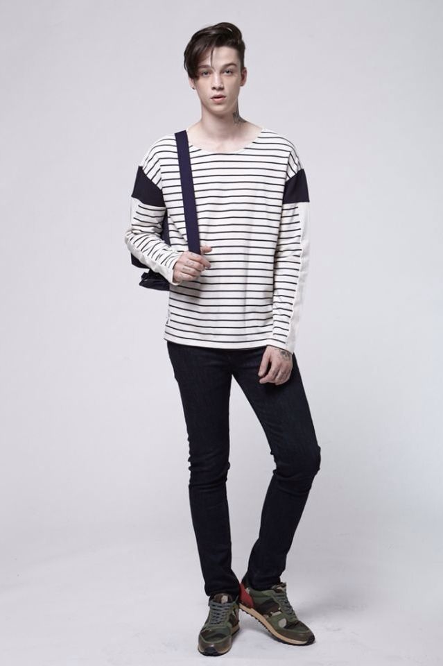 a fullbody studio shot of a male standing in a white room. he is wearing a white and black striped longsleeve tshirt, dark blue jeans, and olive green and red sneakers. he has a black shoulder bag slung over his shoulder.