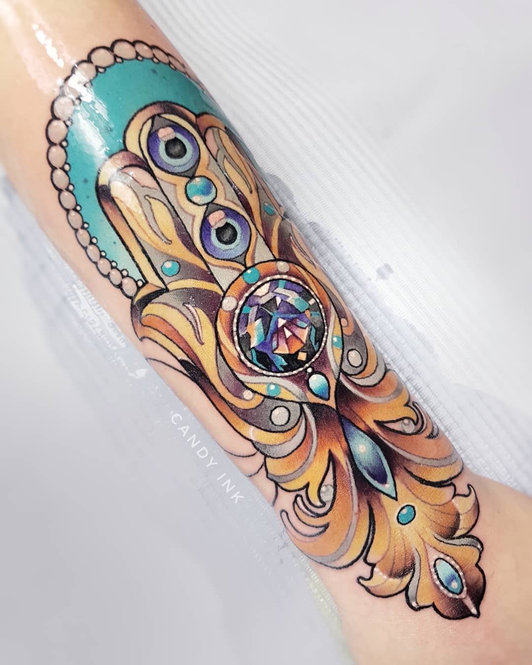 a closeup image showcasing a vibrant tattoo of a hamsa hand on someone's forearm. the hamsa, adorned with ornate patterns and gemstones, takes center stage against a minimalist white background. the tattoo is rendered in a neotraditional style, characterized by bold outlines, rich color gradients, and meticulous detailing. three evil eyes, embedded within the hand's design, add a layer of symbolic depth. the interplay of turquoise, gold, and sapphire hues further accentuates the visual appeal of the tattoo.  the tattoo artist's signature, "candy ink", is subtly incorporated into the artwork.