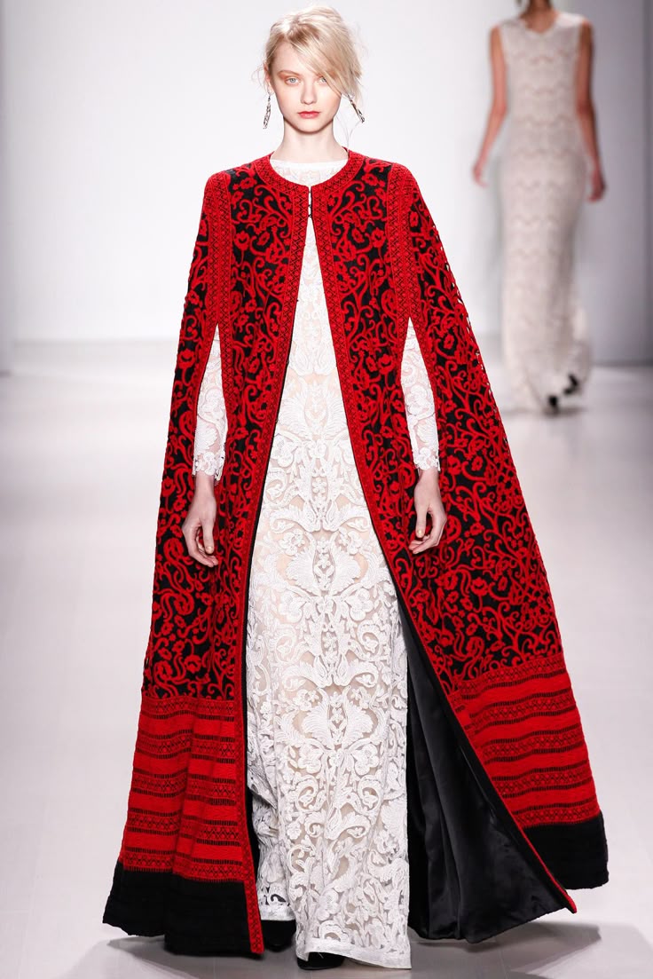 a fashion runway scene featuring a model wearing an elaborate red and black embroidered cape over a white lace gown. another model in a long, white lace dress is walking in the background. the model in the foreground is wearing dangling earrings and has an elegant, highfashion look. the cape has a distinct pattern and reaches down to the floor. the background is a white runway setting.