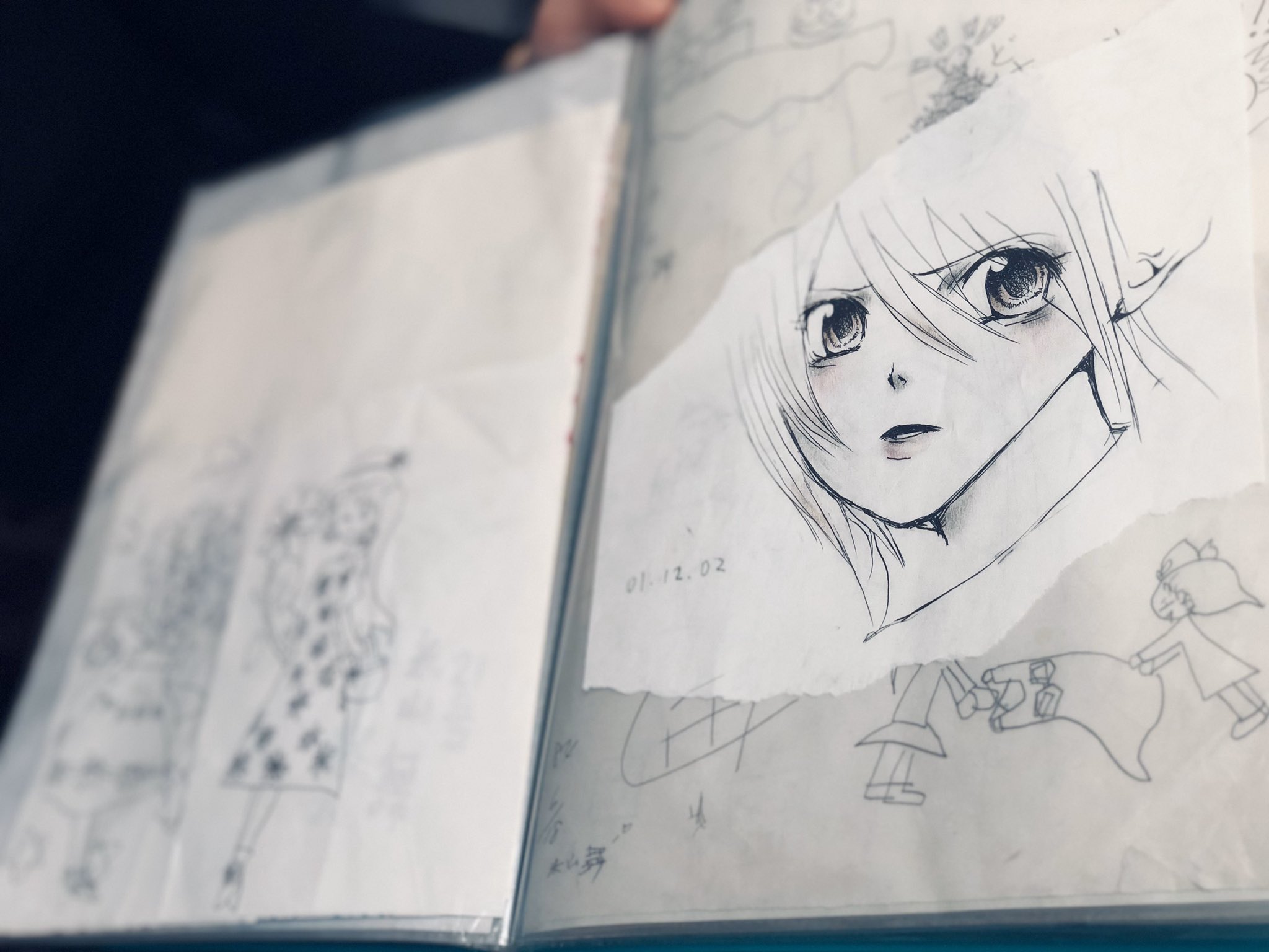 a closeup shot shows an open sketchbook. on the right side, a piece of paper with a black and white animestyle drawing of a girl's head taped onto the sketchbook page. the girl in the drawing has long, white hair, large eyes, and a small mouth. she is looking to the left, and has a slightly worried expression. surrounding the tapedon drawing is another sketch. the left side of the sketchbook shows another, fainter sketch of a figure. the lighting suggests it is daytime, with natural light illuminating the book. the overall impression is of someone working on an anime or manga illustration.